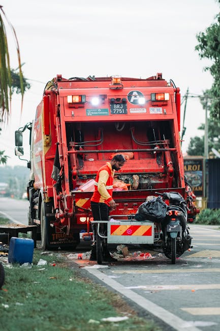 A waste collection worker stands beside a large red rubbish truck on the street, focused on emptying a black trash bag into the vehicle's rear opening. The worker wears a high-visibility yellow and orange vest over dark trousers, with gloves on the hands. The truck, branded with the company logo, has a metallic, slightly weathered finish with reflective orange and yellow safety markings on the back. The scene is set on a paved urban road with a curb and a patch of green grass nearby, and a motorcycle parked adjacent to the truck. In the background, there are blurred trees, utility poles, and a building with signage, indicating a commercial or mixed-use area. Overcast daylight conditions create soft, diffuse lighting across the scene, emphasizing the practical and service-oriented nature of waste collection operations, which are a key part of alternative waste handling and rubbish removal services provided by Waste Collection Brent.