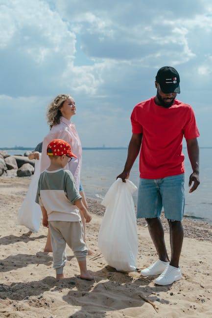 A family participating in a beach clean-up activity by collecting litter with large white trash bags. The scene takes place on a sandy shore near a body of water, with a rocky area visible in the background and a partly cloudy sky overhead. The father, dressed in a red t-shirt, denim shorts, and a black cap, is holding a white trash bag and appears to be engaged in gathering debris. A woman, wearing a light pink top, stands nearby observing, while a young boy dressed in a grey t-shirt, beige shorts, and a red cap with yellow patterns is also holding a trash bag, ready to assist. The environment reflects a casual, outdoor setting focused on environmental cleanliness, which aligns with the principles of private waste handling and rubbish removal services provided by Waste Collection Brent, especially in the context of alternative waste collection efforts outside municipal services.