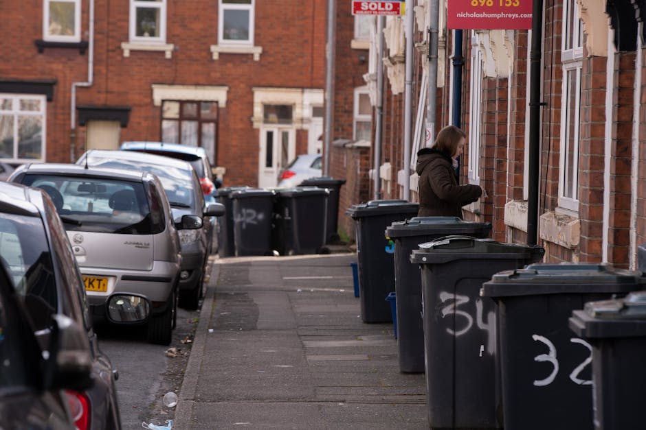 A row of large, black wheelie bins with closed lids are lined up along the edge of a residential pavement outside a series of red brick terraced houses. The bins are positioned close to the front walls of the buildings, which feature white window frames and decorative stone lintels. In the background, a woman dressed in a dark coat with a hood is seen standing next to one of the bins, appearing to be disposing of waste. Several parked cars are visible on the street, including a silver hatchback in the foreground with a visible yellow registration plate. The street is relatively narrow, and the scene is illuminated by natural daylight, with subtle reflections on the vehicle surfaces. The environment suggests a typical urban residential area where private waste collection services, such as those provided by Waste Collection Brent, may be used for rubbish disposal and on-site clearance, as evidenced by the multitude of bins designated for different waste types or collection days.